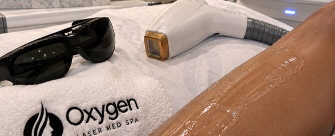 Laser hair removal treatment prep at Oxygen Med Spa, showing cooling gel applied to a client's leg with the laser device and protective eyewear nearby