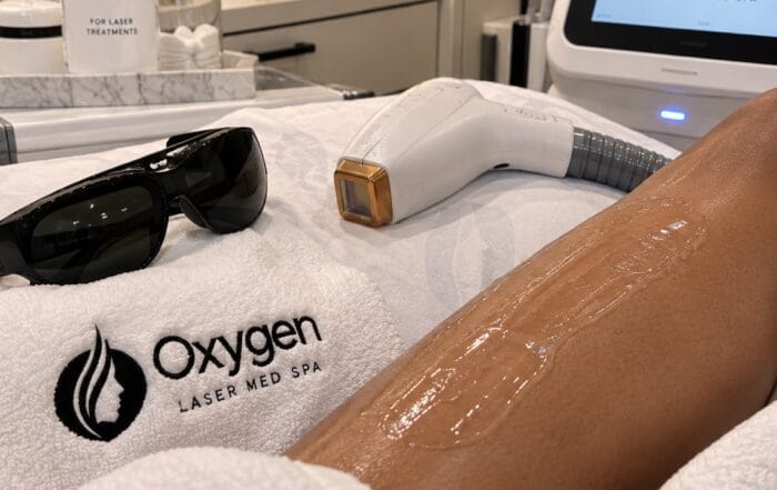 Laser hair removal treatment prep at Oxygen Med Spa, showing cooling gel applied to a client's leg with the laser device and protective eyewear nearby